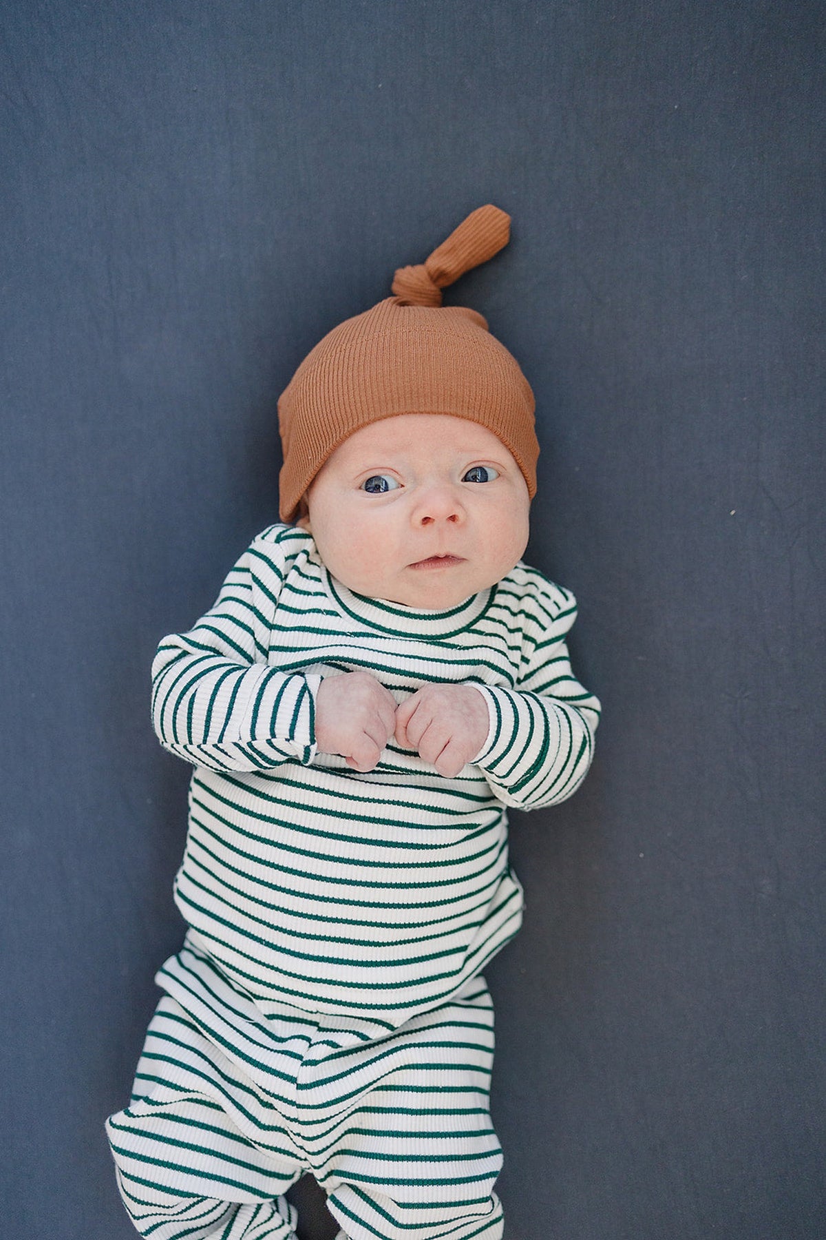 Baby in a striped outfit with a brown hat lying on a dark charcoal crib sheet.