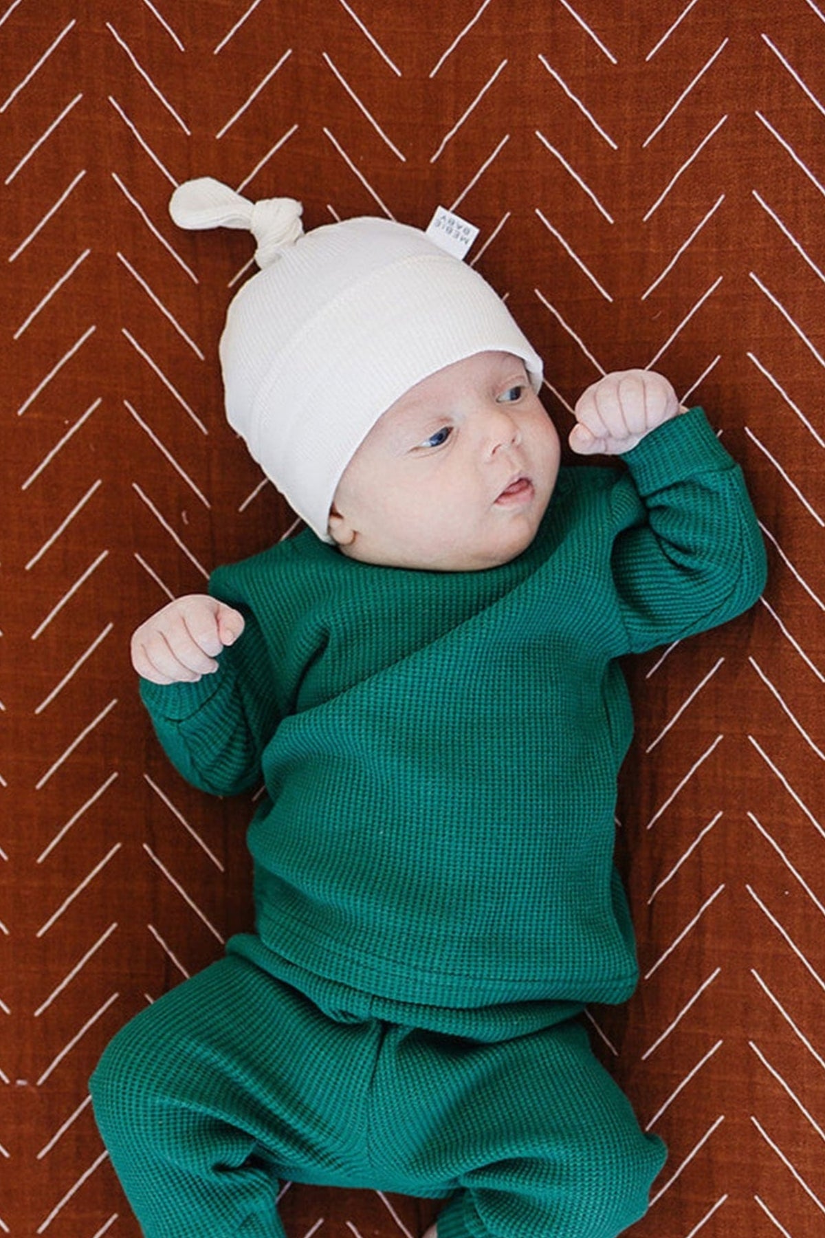 Newborn wearing a white knot hat and green outfit lies on a patterned brown blanket.