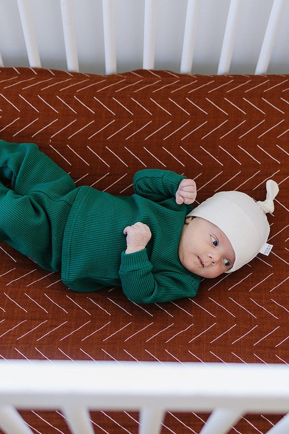 A baby in green clothing and a white hat lies on a rust-colored, patterned crib sheet.