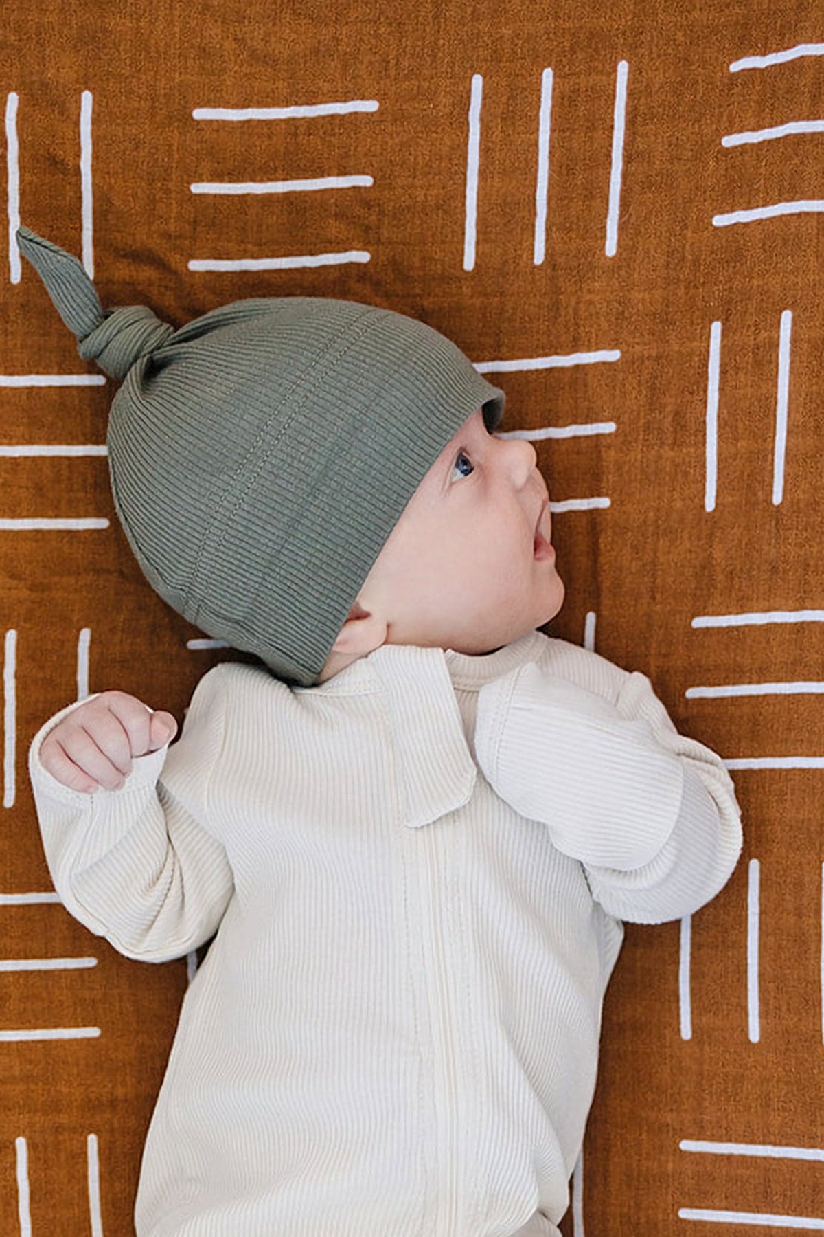 A baby wearing a green ribbed knot hat and cream onesie lies on a brown patterned blanket.