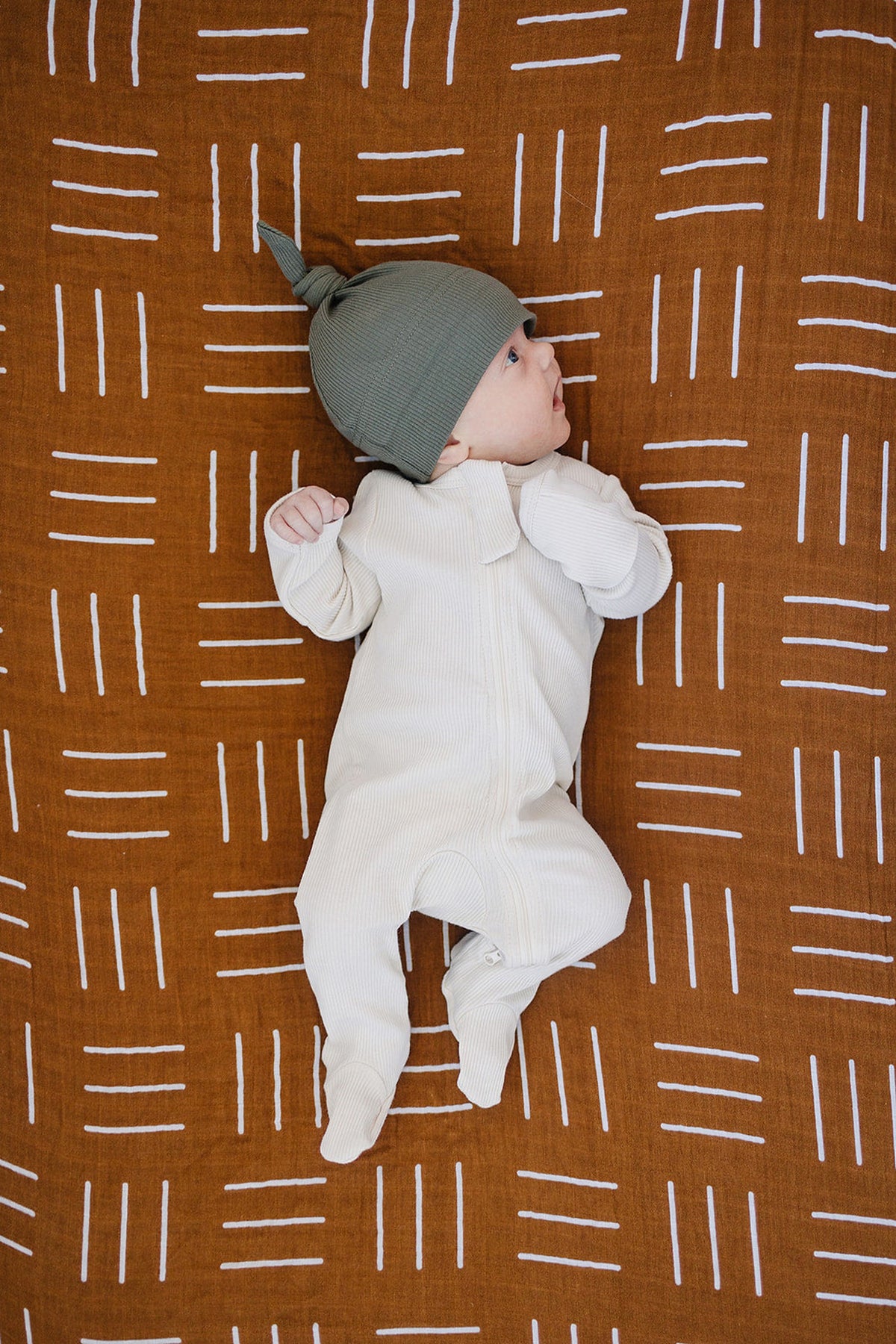 A baby in a cream outfit and green hat lies on a mustard mudcloth muslin crib sheet with white patterns.