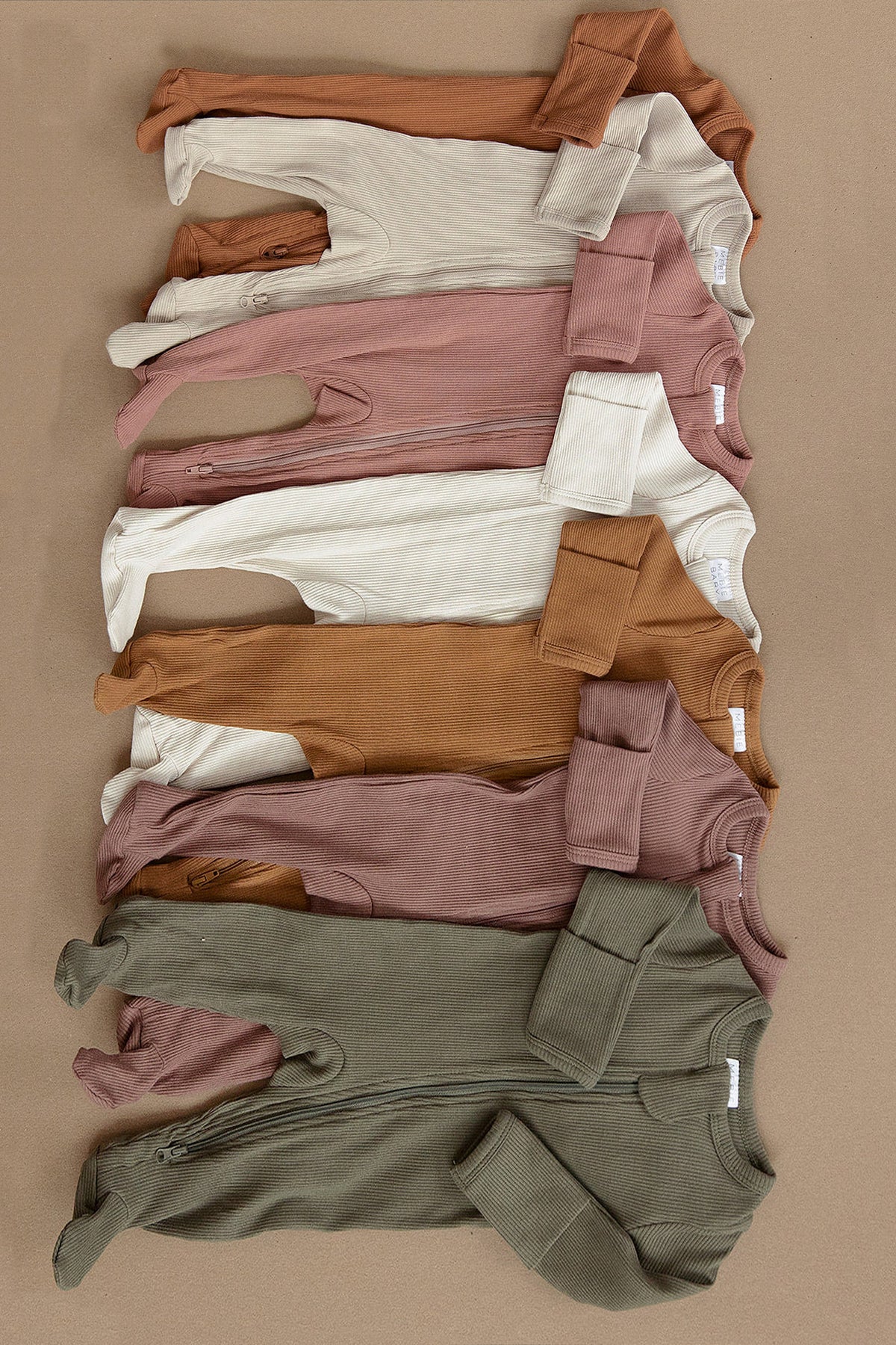 A variety of ribbed zipper footed pajamas in earth tones, neatly arranged on a beige background.