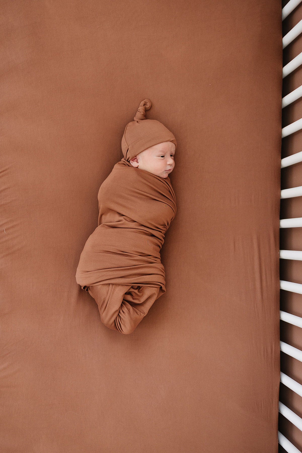 A baby wrapped in a rust-colored swaddle and hat lies on a matching crib sheet with white bars.