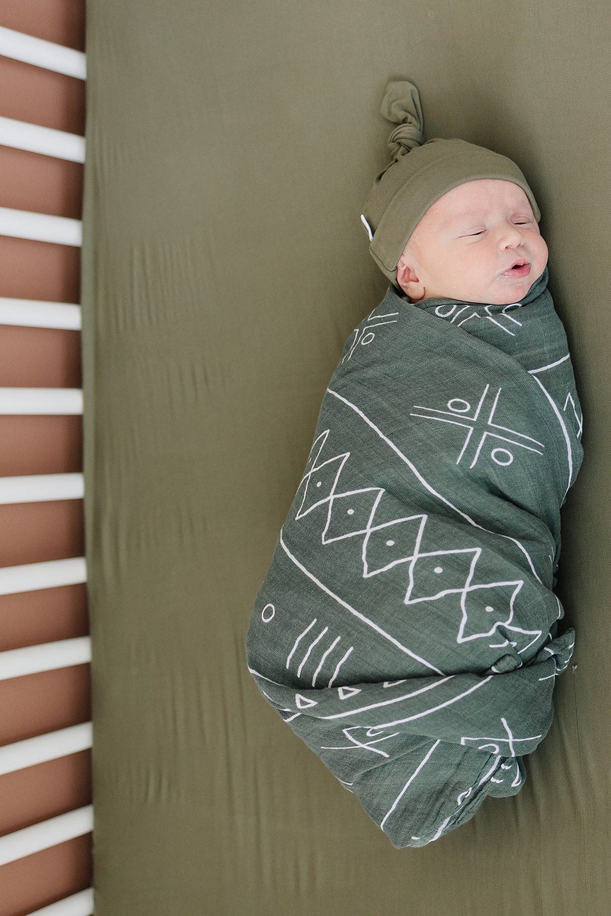 A baby is swaddled in a patterned green blanket, lying on an olive crib sheet with wooden slats visible.