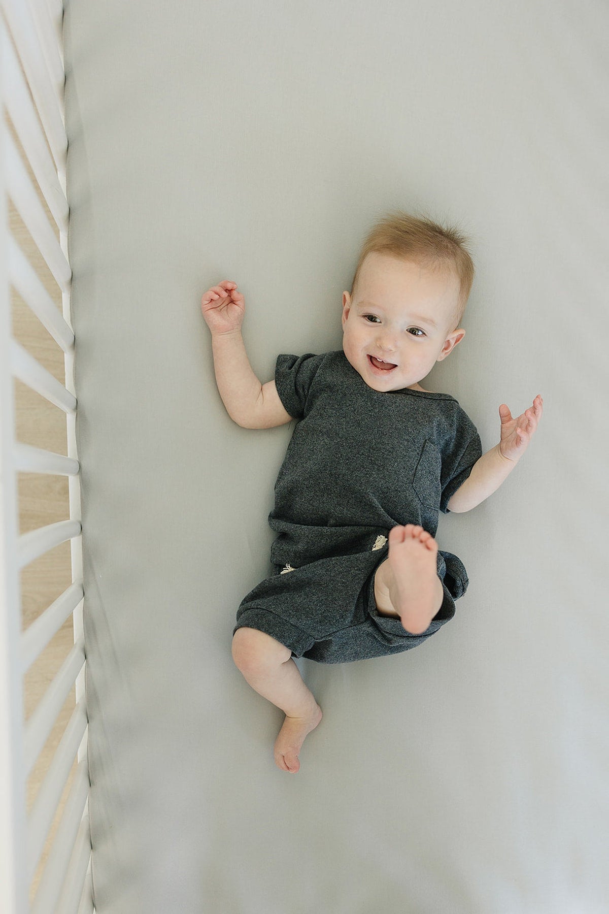 Smiling baby in a dark gray outfit lies on a light gray crib sheet inside a white crib.