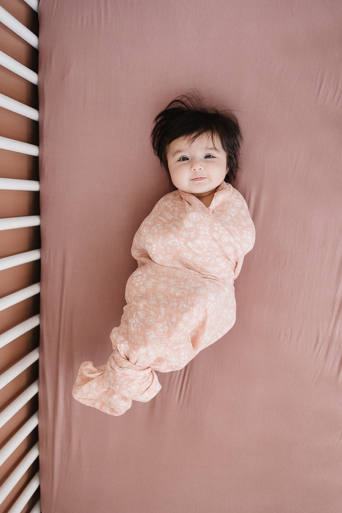 A baby is swaddled in a floral patterned blanket on a dusty rose crib sheet.