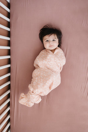 A baby is swaddled in a floral patterned blanket on a dusty rose crib sheet.