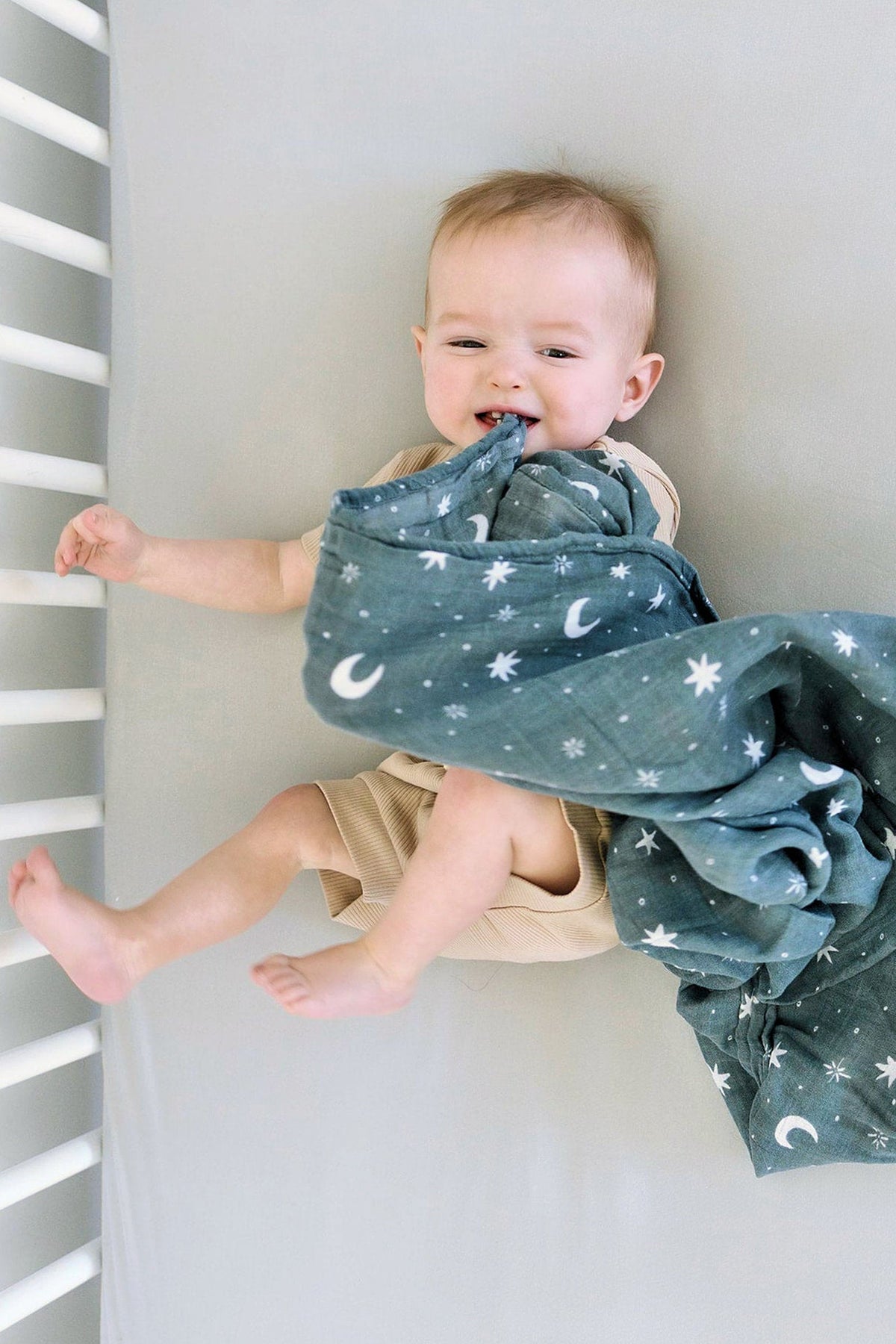 A baby lies on a light-colored crib sheet, playfully chewing a blue blanket with moon and star patterns.