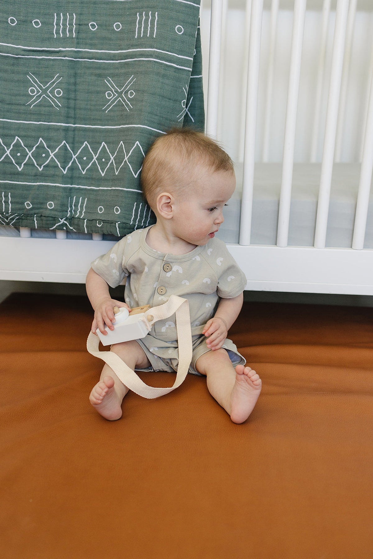 A baby in patterned clothing holds a toy camera, sitting on a brown surface beside a crib with a green.