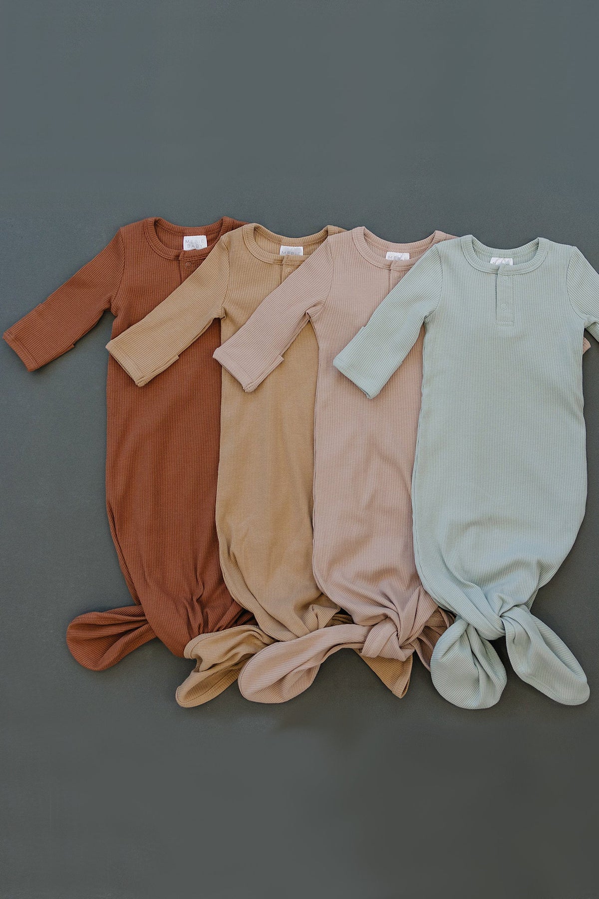 Four ribbed knot baby gowns in brown, beige, pink, and sage green are neatly arranged on a gray background.
