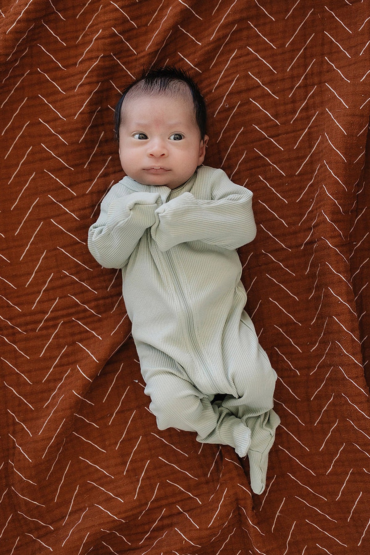 A baby lies on a patterned brown blanket, wearing sage ribbed zipper footed pajamas made from organic cotton.