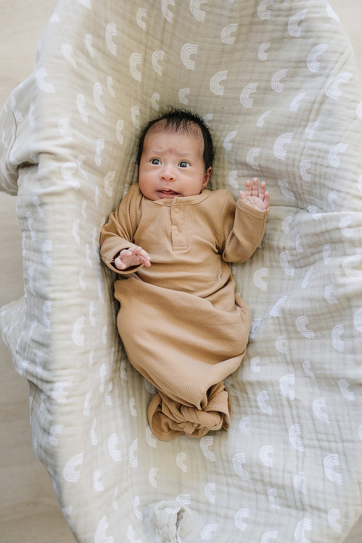 A baby wearing a tan ribbed gown lies on a patterned blanket. The gown is made of organic cotton.