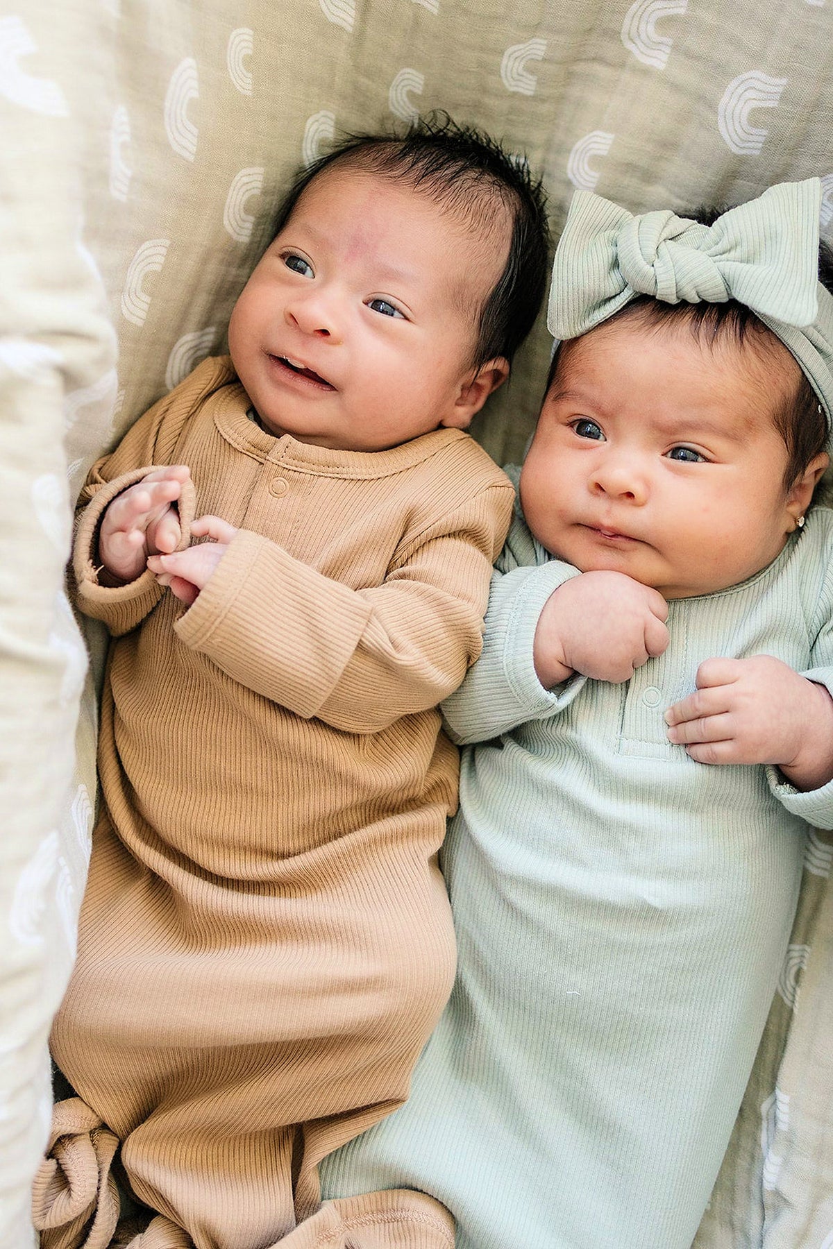 Two babies lie on a patterned blanket, wearing ribbed gowns; one in beige, the other in light green.