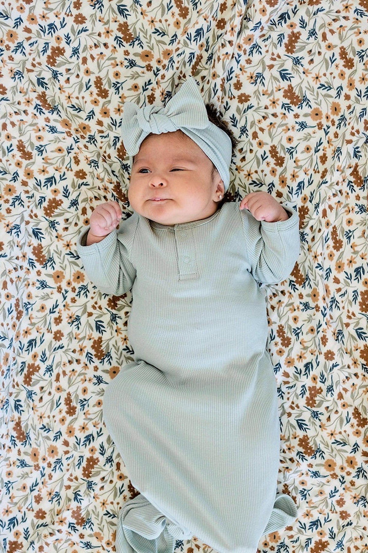 A baby in a sage ribbed outfit and matching head wrap lies on a floral-patterned blanket.