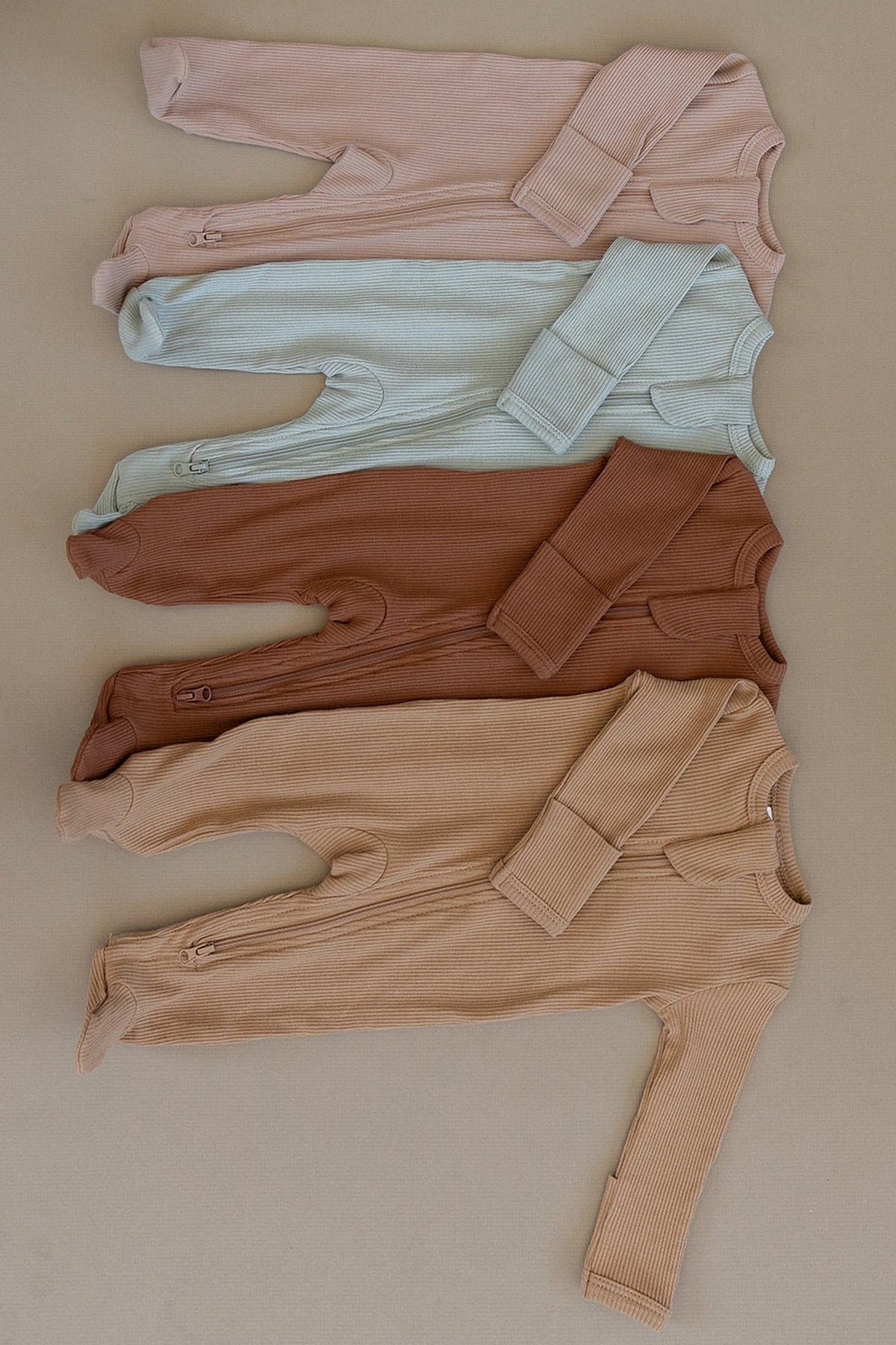 Four ribbed zipper footed baby pajamas in pale pink, light blue, brown, and beige, laid flat on a surface.