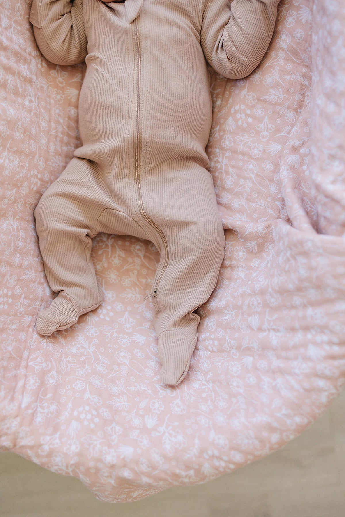 A baby wearing a pale pink ribbed zipper footed pajama lies on a floral-patterned pink blanket.