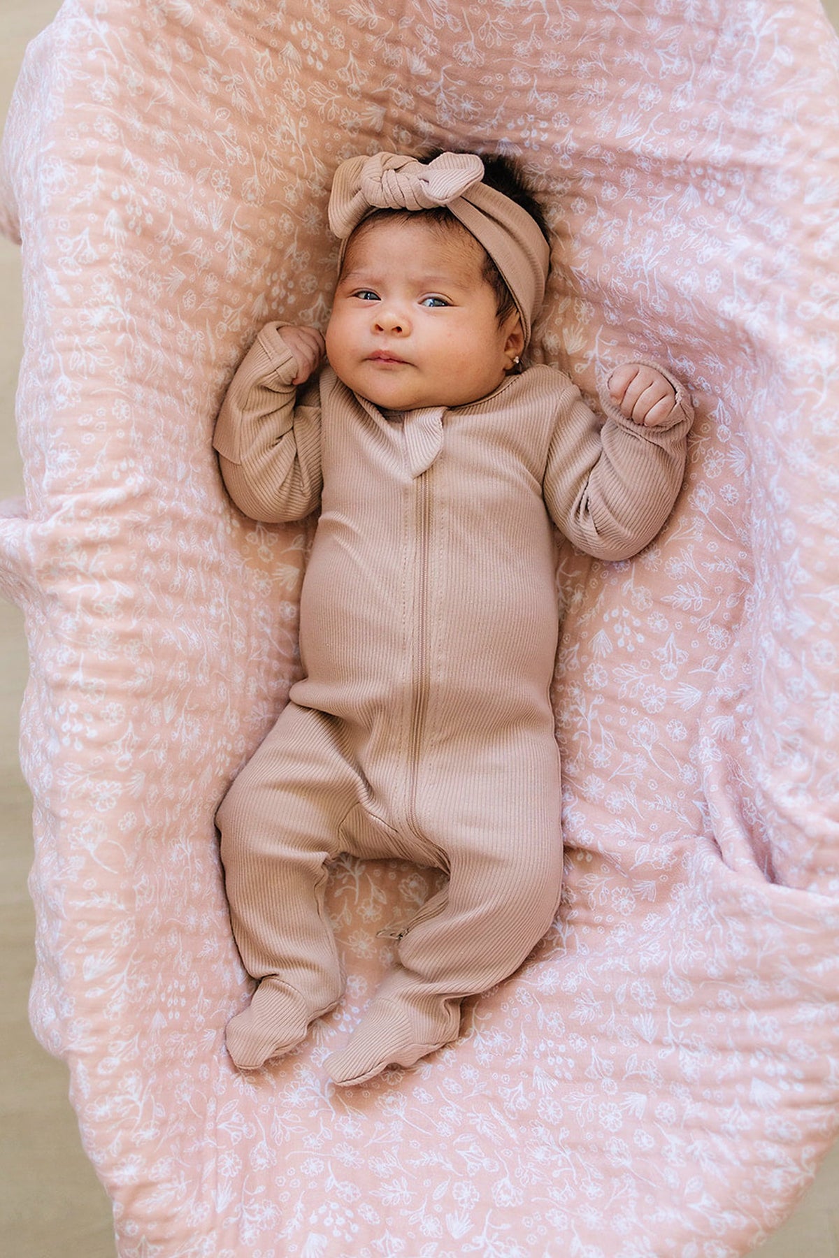 A baby in a pale pink ribbed zipper footed pajama with a matching headband lies on a floral blanket.