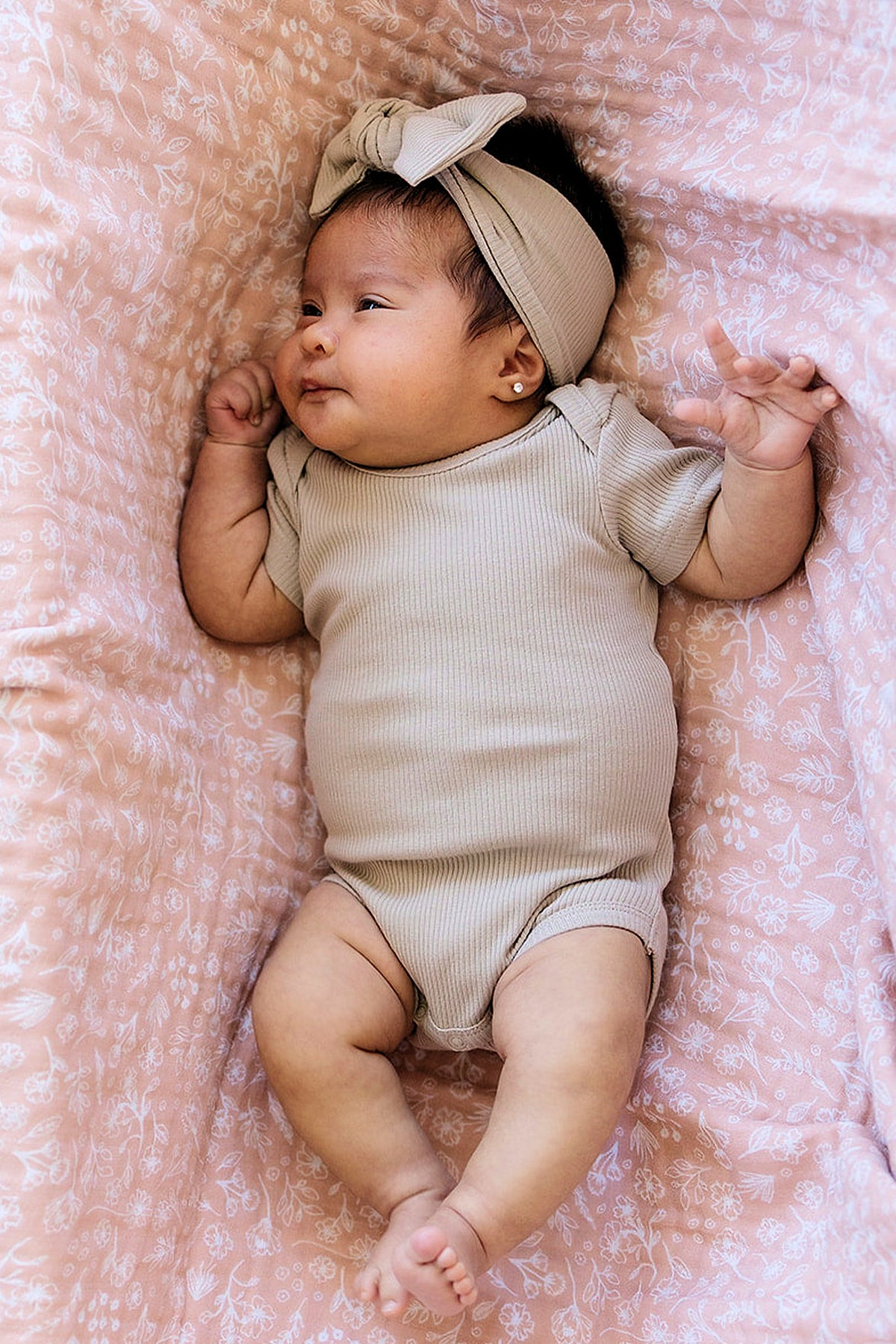 A baby wearing an oatmeal ribbed onesie and head wrap lies on a pink floral-patterned blanket.