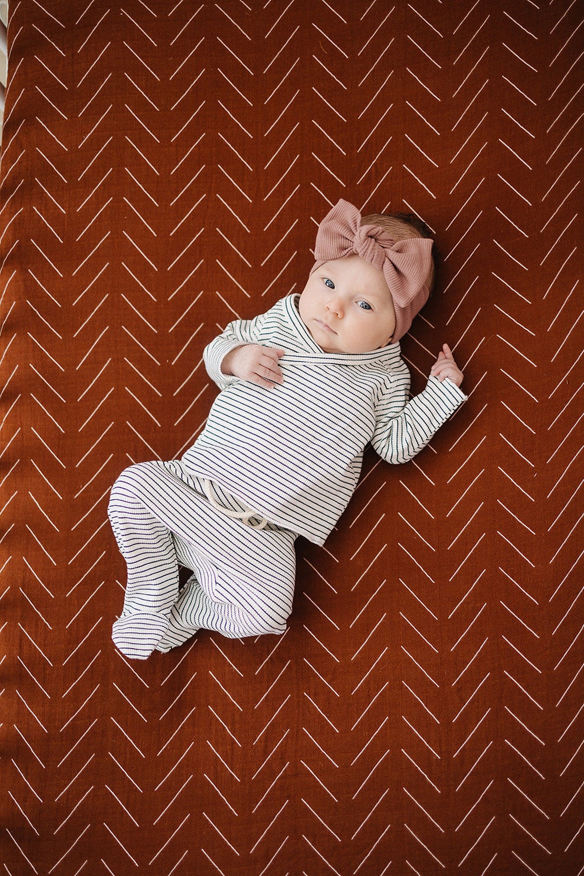 A baby lies on a rust-colored crib sheet with a white geometric pattern, wearing a striped outfit and bow.