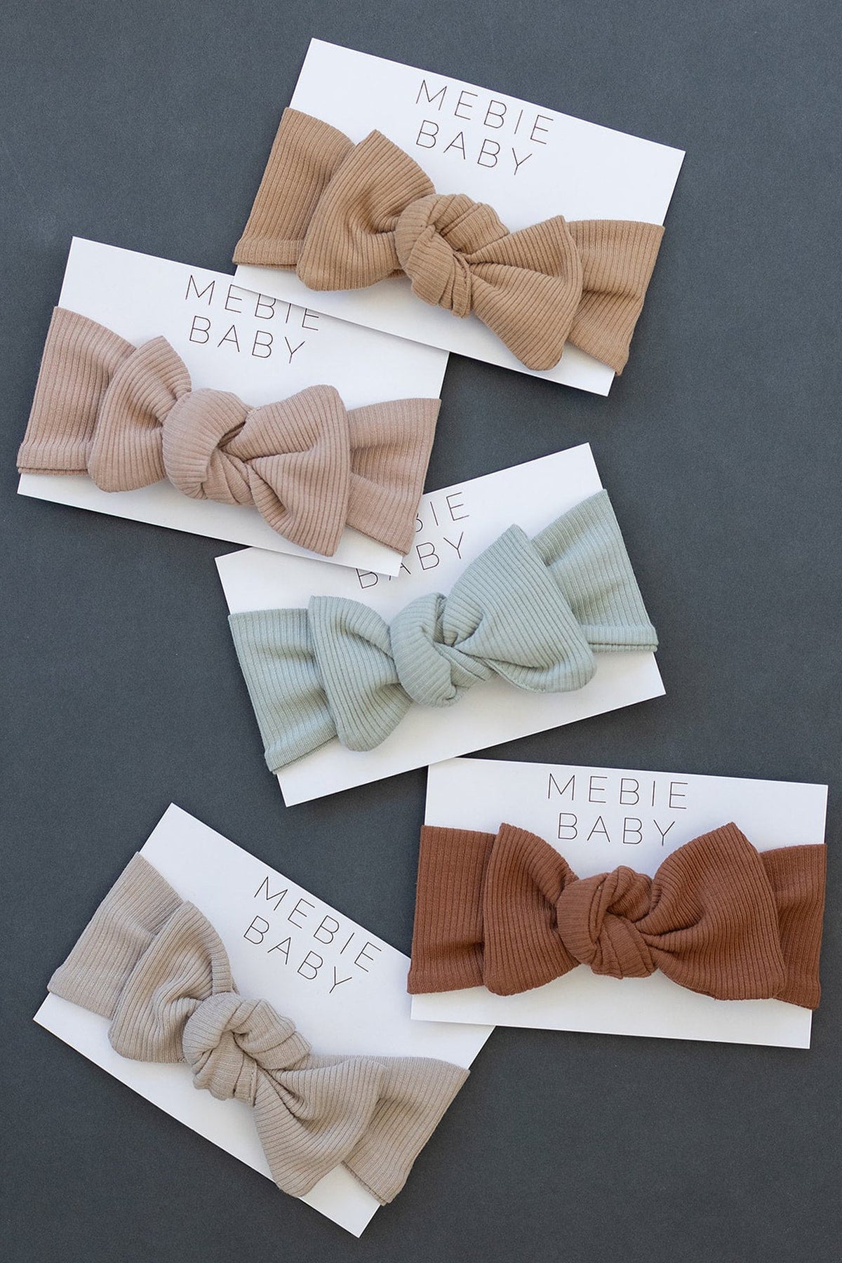 Five ribbed head wraps in neutral tones are displayed on Mebie Baby cards, showcasing simple square knot designs.