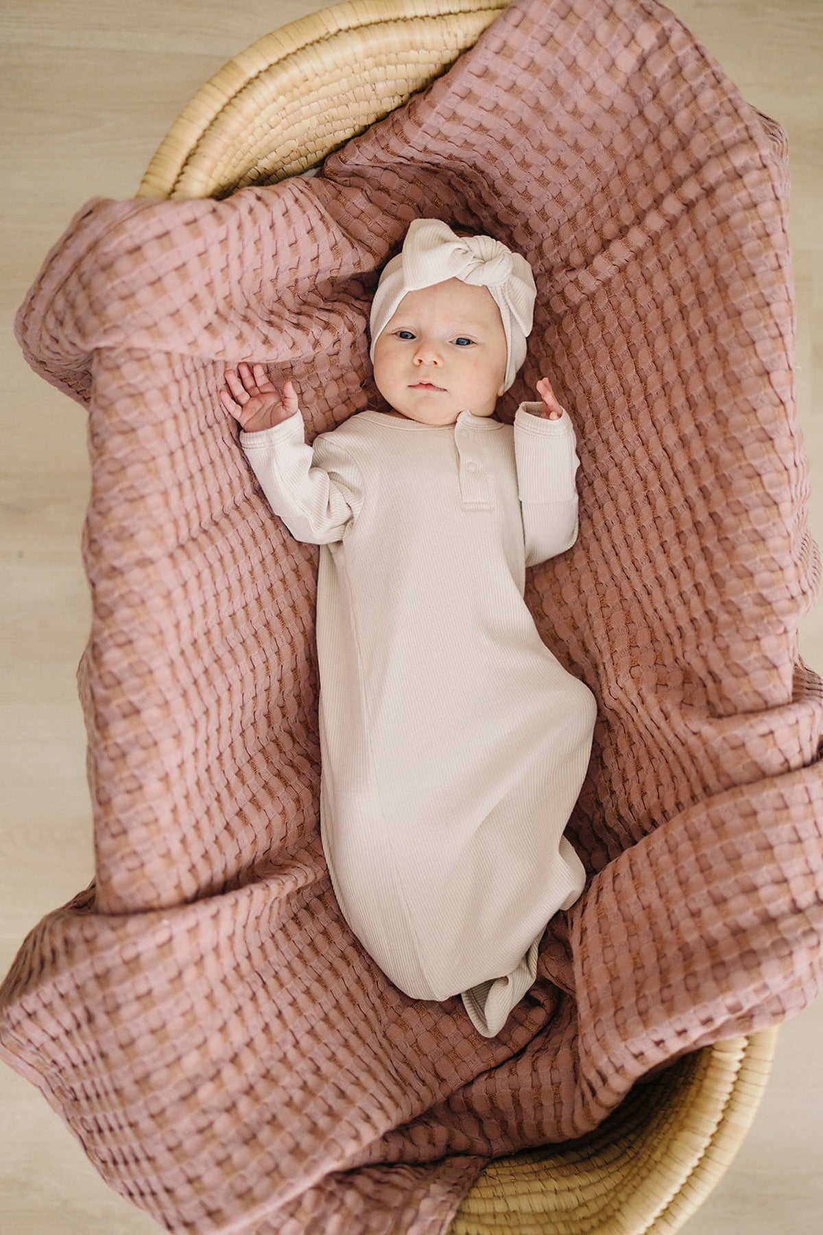 A baby wearing a vanilla ribbed knot gown and headband lies in a basket with a pink textured blanket.