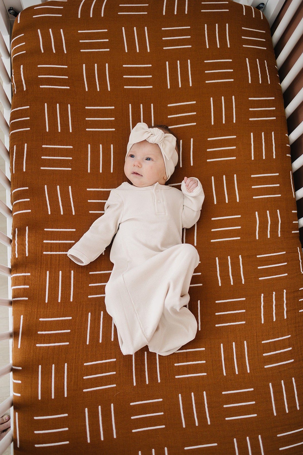 A baby in a cream outfit and headband lies on a mustard mudcloth patterned crib sheet in a white crib.