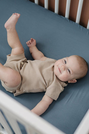 A baby in a beige outfit lies on a dusty blue crib sheet in a white crib.