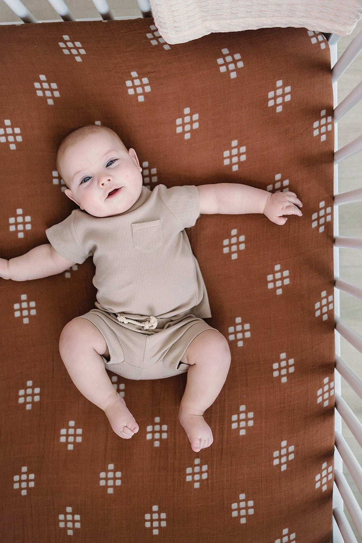 A baby lies on a brown muslin crib sheet with a white geometric pattern, wearing a beige outfit.