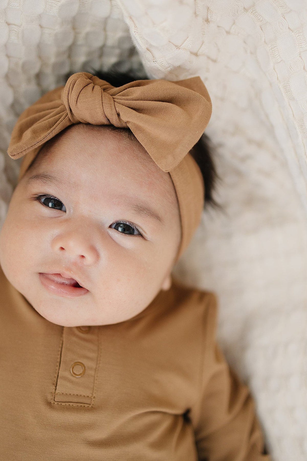 Baby wearing a mustard head wrap tied in a bow, lying on a textured cream blanket.