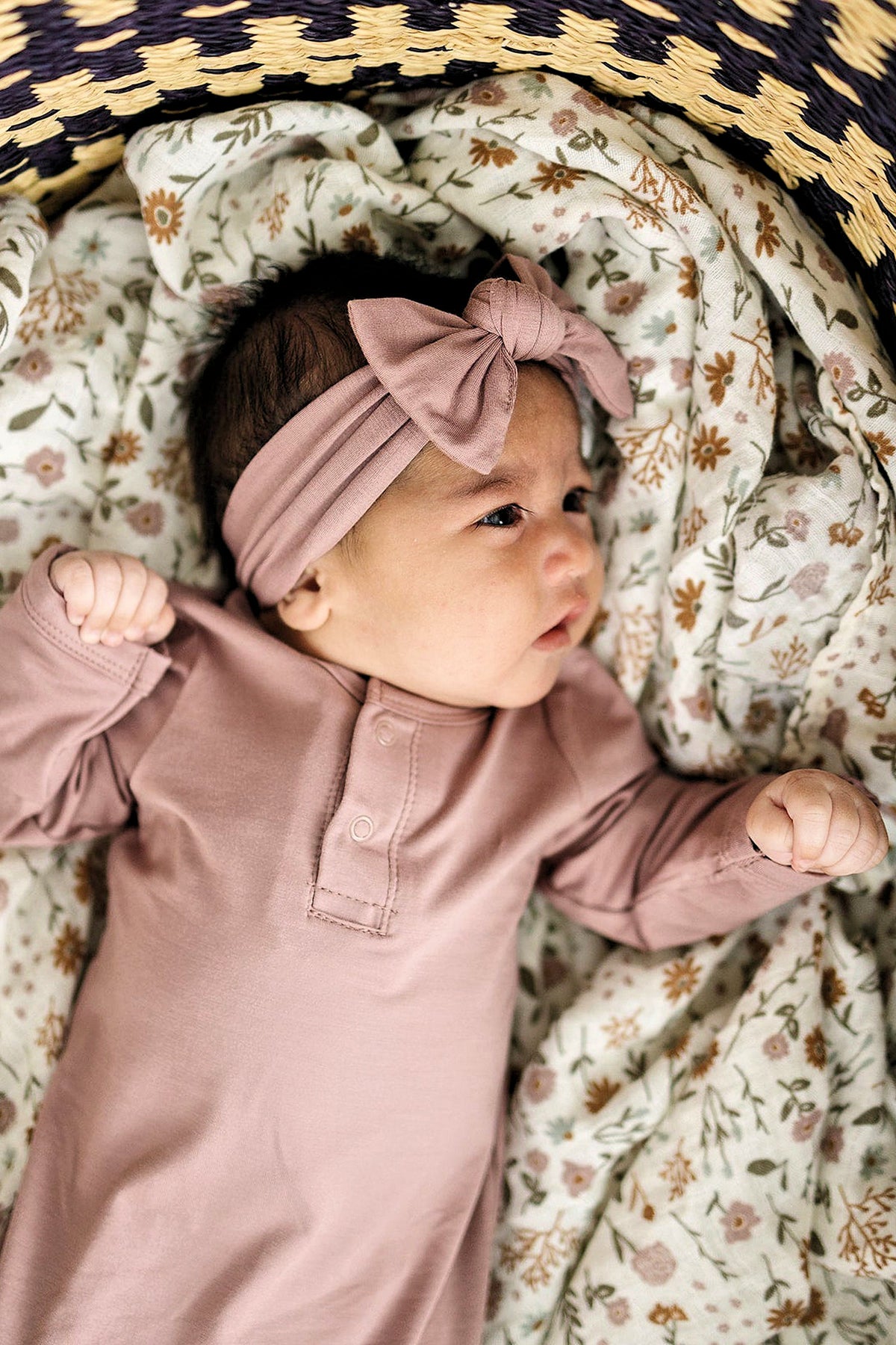 A baby wearing a dusty rose head wrap and gown lies on a floral-patterned blanket in a woven basket.