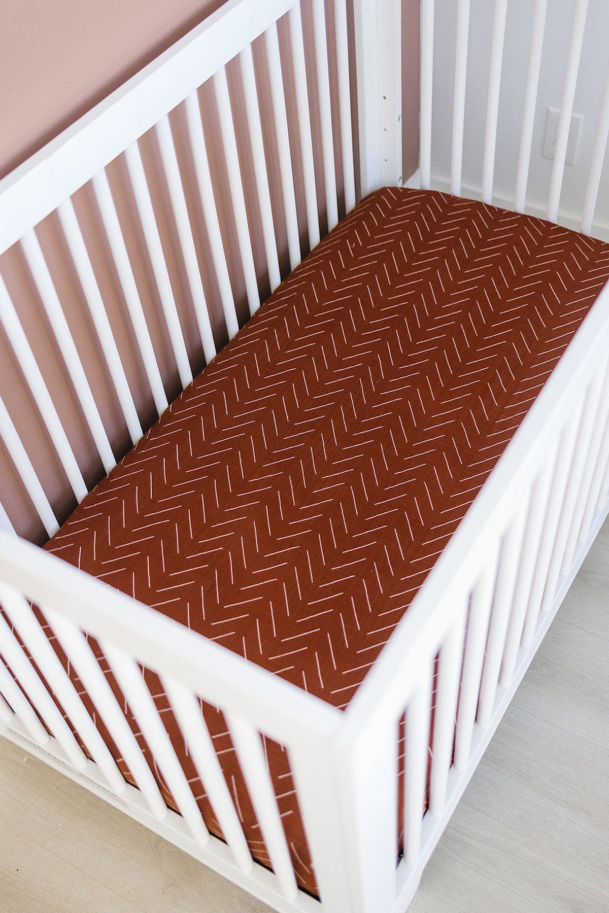White crib with a rust-colored mudcloth muslin crib sheet featuring a subtle zigzag pattern.