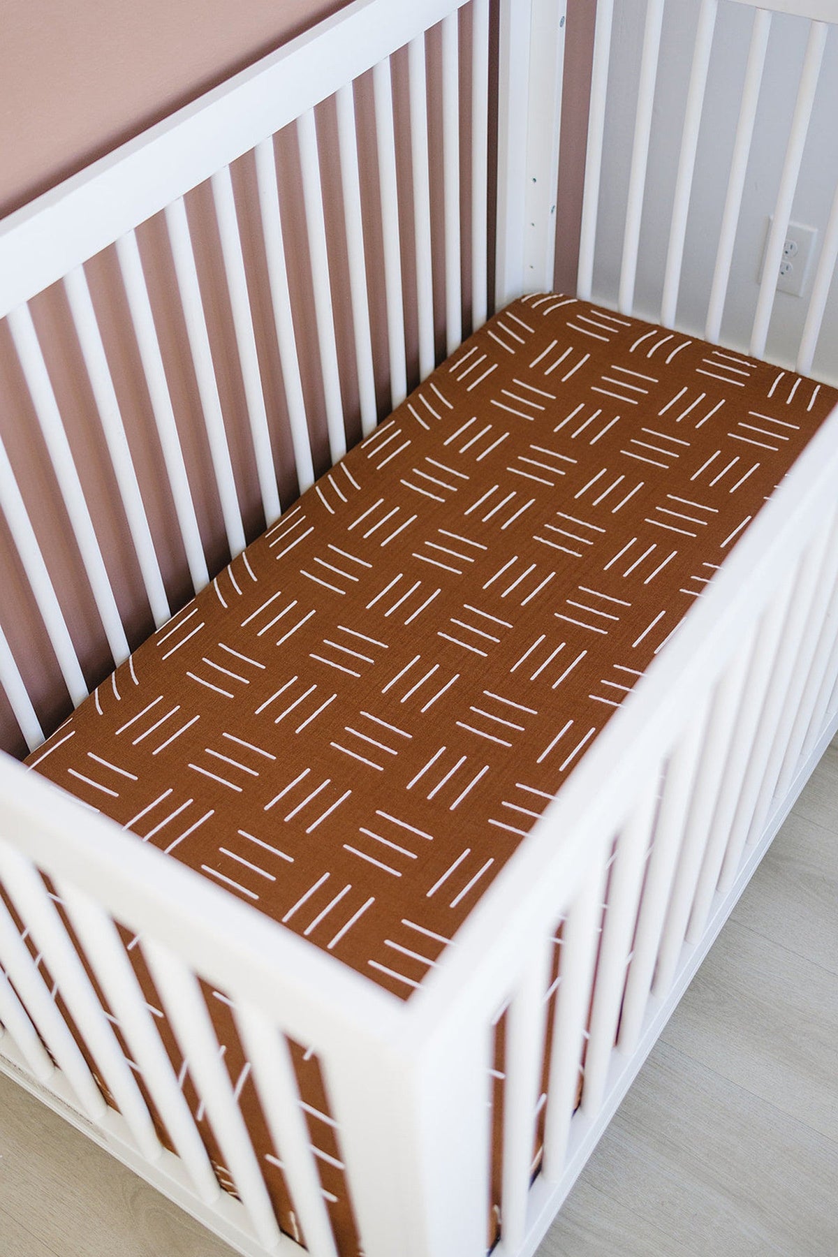 White crib with a mustard mudcloth muslin sheet featuring a geometric pattern, set against a light wooden floor.