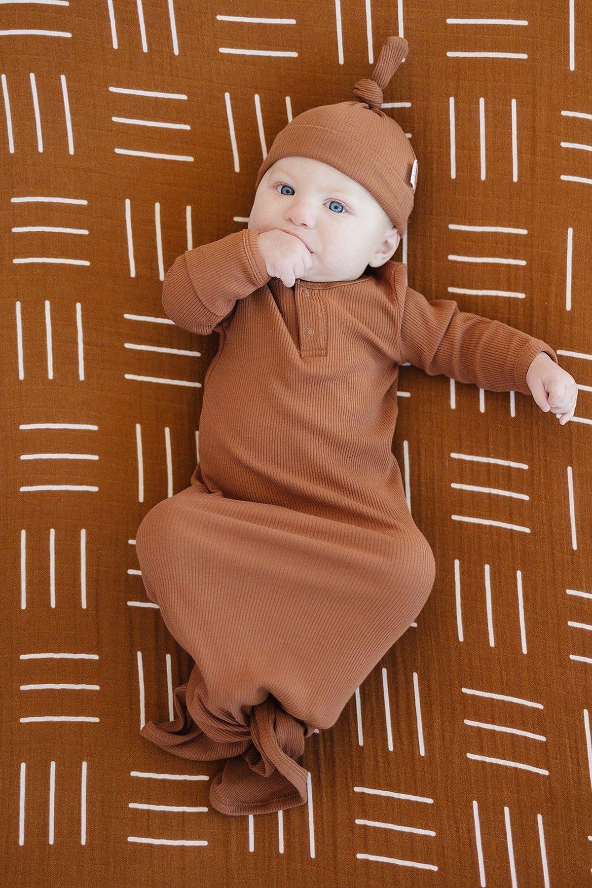 A baby in a brown outfit lies on a mustard mudcloth patterned crib sheet, featuring white geometric lines.