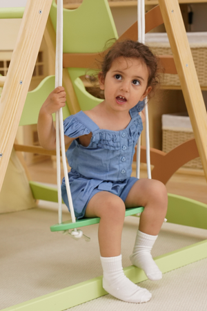 A young girl with curly hair sits on a green swing, wearing a light blue romper with ruffles. The background features a wooden jungle gym in a bright playroom setting.