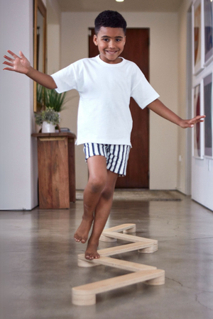 A young boy balances on a wooden balance beam in a modern interior space. He wears a white t-shirt and striped shorts, showcasing coordination and confidence during playtime.