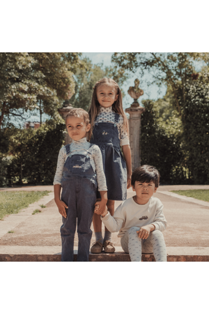 Three children stand outdoors on a stone pathway, wearing stylish outfits: two in denim overalls and one in a light long-sleeve shirt, all showcasing a playful, casual look.