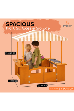 Children play together at a wooden outdoor market station with a striped canopy. The setup features spacious work surfaces and storage, designed for ages three and up.