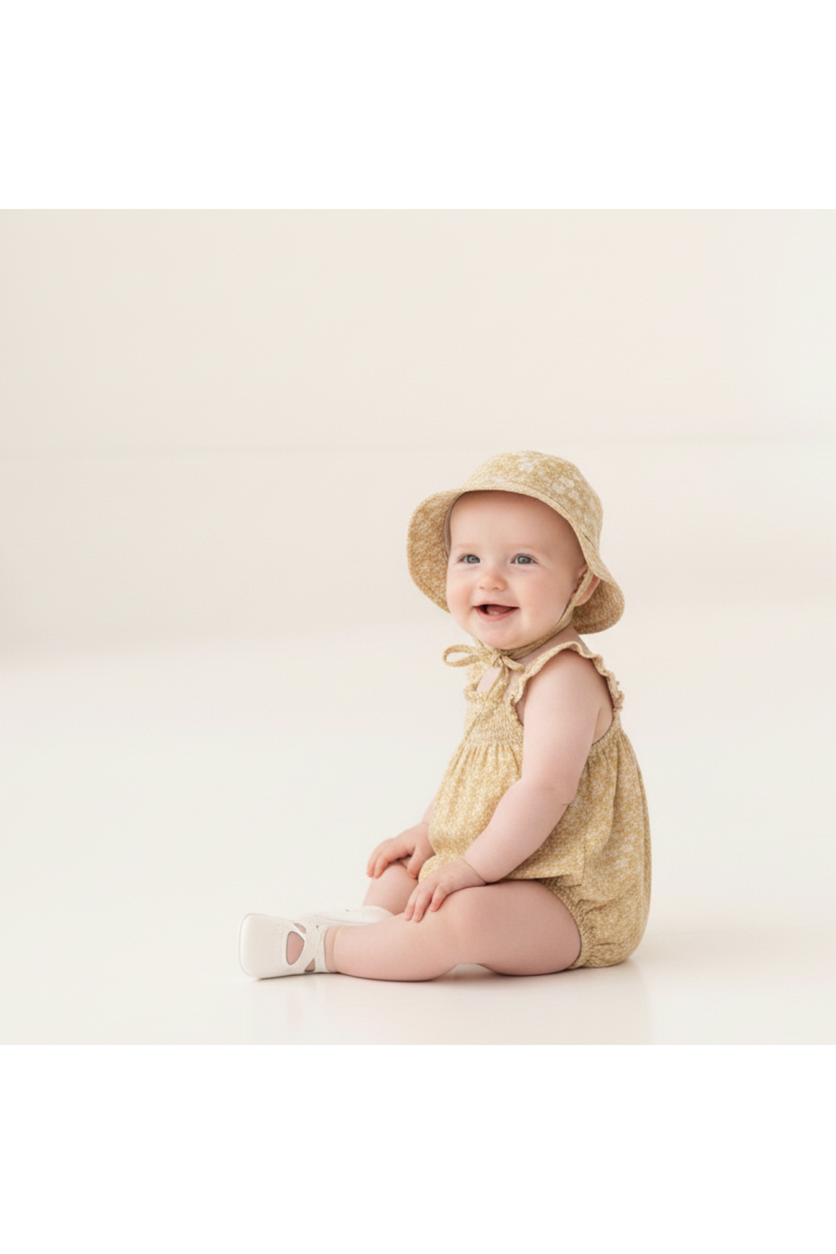 A smiling baby sits on a light, smooth surface, wearing a yellow floral bubble romper with ruffle sleeves and a matching sun hat.