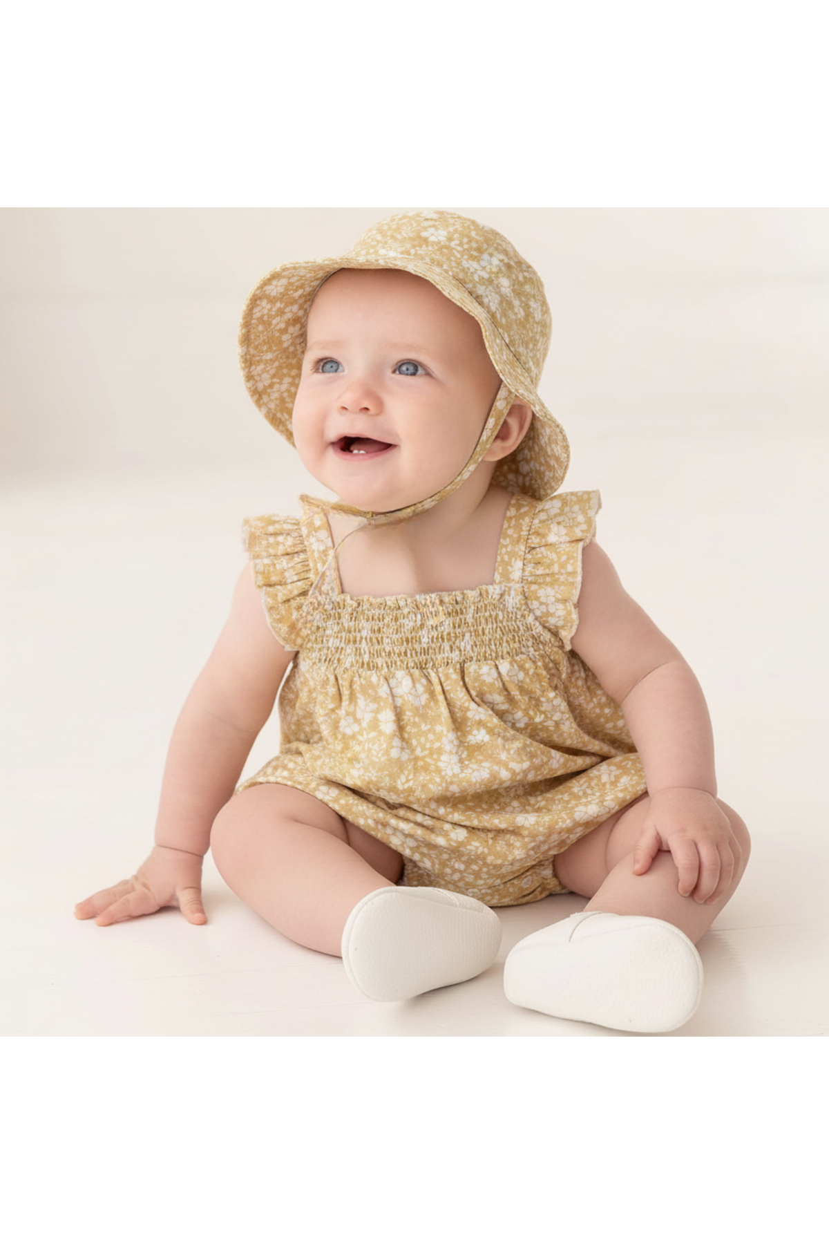 A cheerful baby sits on a light background, wearing a yellow floral ruffle sleeve bubble romper and matching sun hat. The outfit is made from soft, organic cotton for comfort.