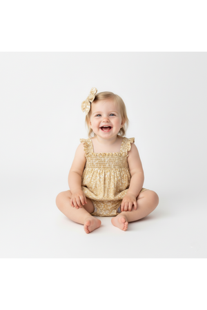 A joyful toddler sits on a light background, wearing a soft yellow bubble romper with ruffle sleeves and a floral pattern.