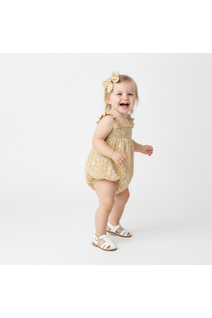 A joyful toddler wears a yellow floral ruffle sleeve bubble romper, paired with white sandals. The organic cotton fabric ensures comfort and breathability for active play.