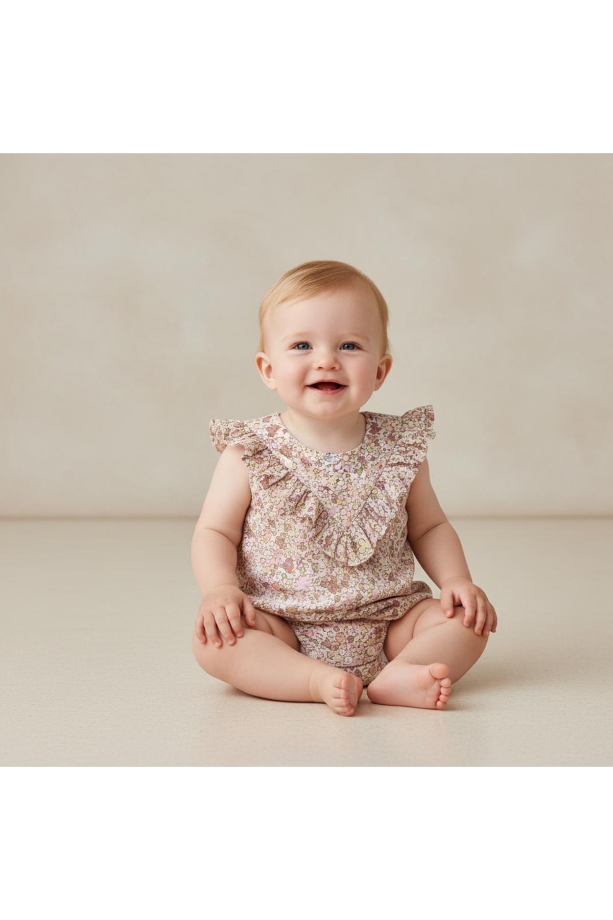 A smiling baby sits on a soft surface, wearing a pastel floral sleeveless romper with ruffles. The outfit is made from organic cotton, perfect for comfort and play.