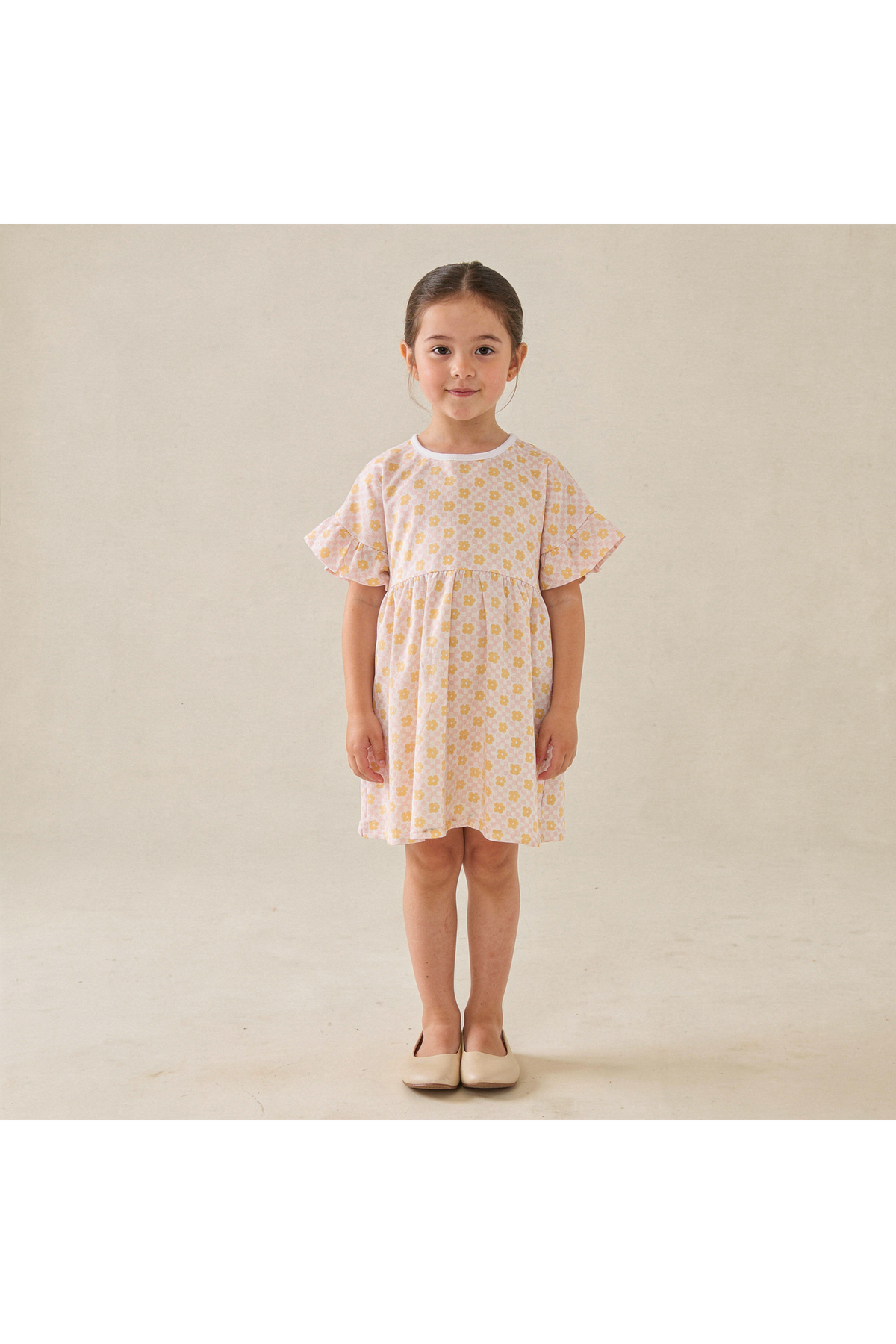 A young girl stands wearing a short-sleeve, organic cotton dress in a soft yellow with a floral print. The dress features a relaxed fit and gently flares at the sleeves.