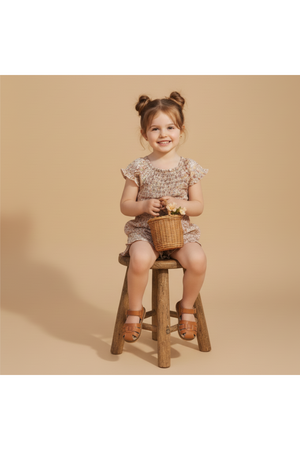 A smiling young girl sits on a rustic wooden stool, wearing a pastel floral smocked jumpsuit. She holds a small woven basket, showcasing a playful, charming look.