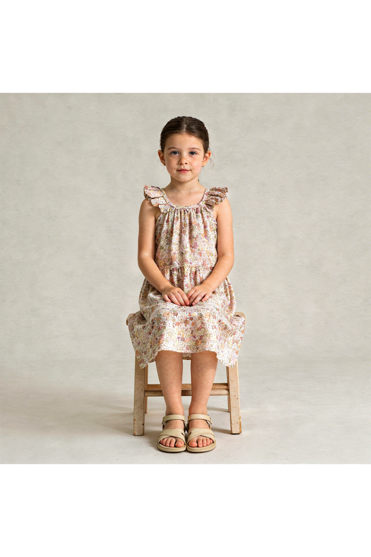 A young girl sits on a wooden stool, wearing a pastel floral ruffle tiered dress made from organic cotton. The dress features a relaxed fit and delicate ruffle details.