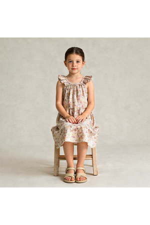 A young girl sits on a wooden stool, wearing a pastel floral ruffle tiered dress made from organic cotton. The dress features a relaxed fit and delicate ruffle details.