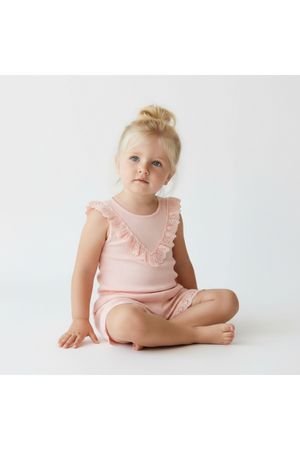 A young child sits on a white background, wearing a soft blush ruffle top and matching shorts set made from organic fabric.