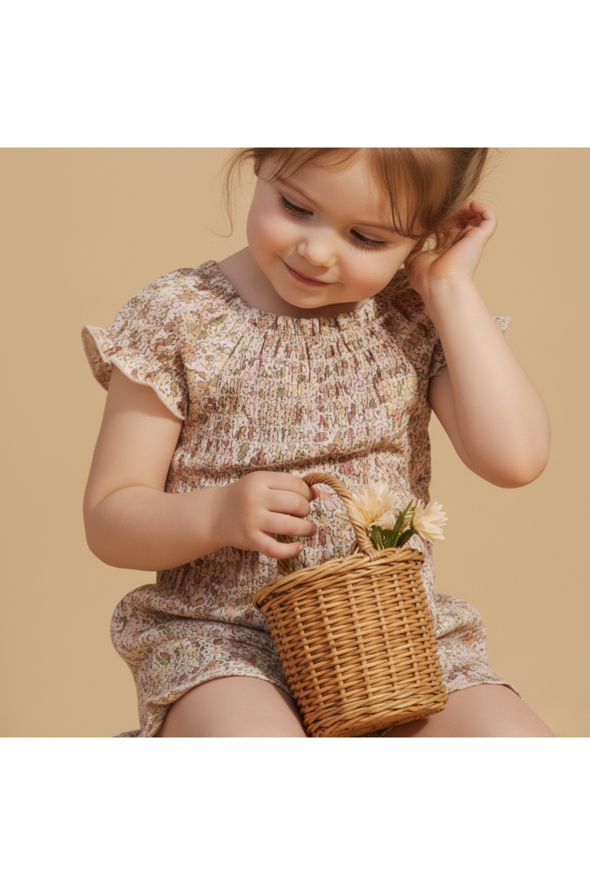 A young child smiles while holding a small wicker basket filled with flowers. She wears a pastel floral smocked jumpsuit.