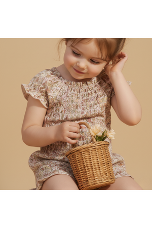 A young child smiles while holding a small wicker basket filled with flowers. She wears a pastel floral smocked jumpsuit.