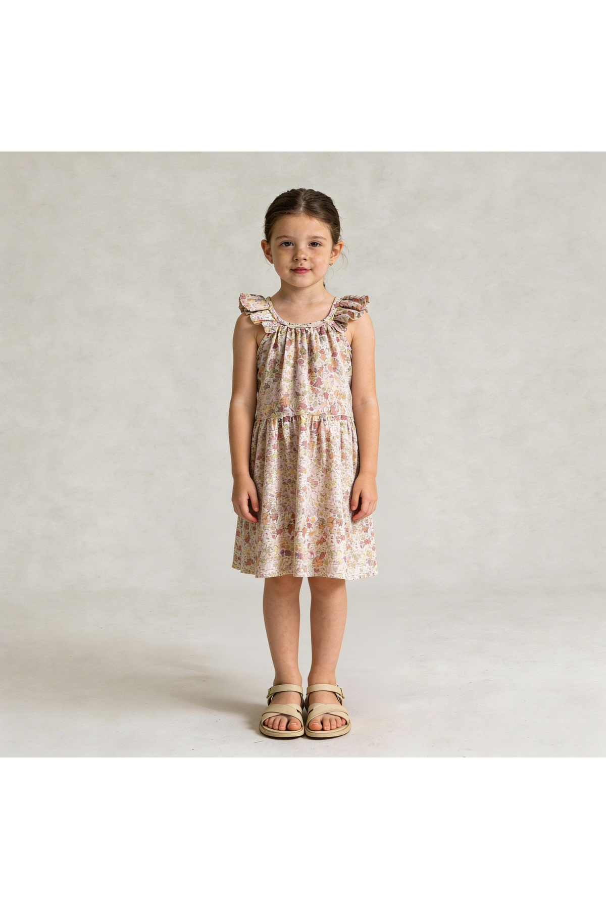 A young girl stands wearing a pastel floral ruffle tiered dress, showcasing a relaxed silhouette and delicate ruffles. The dress is made from organic materials, perfect for sunny days.